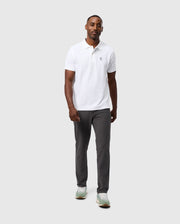 A man stands against a gray backdrop in a white collared short-sleeve shirt and Psycho Bunny MENS MAVERICK 10K PANT - B6P788D200, notable for their five-pocket design. He matches them with light green and white sneakers. His expression is neutral, arms relaxed at his sides.