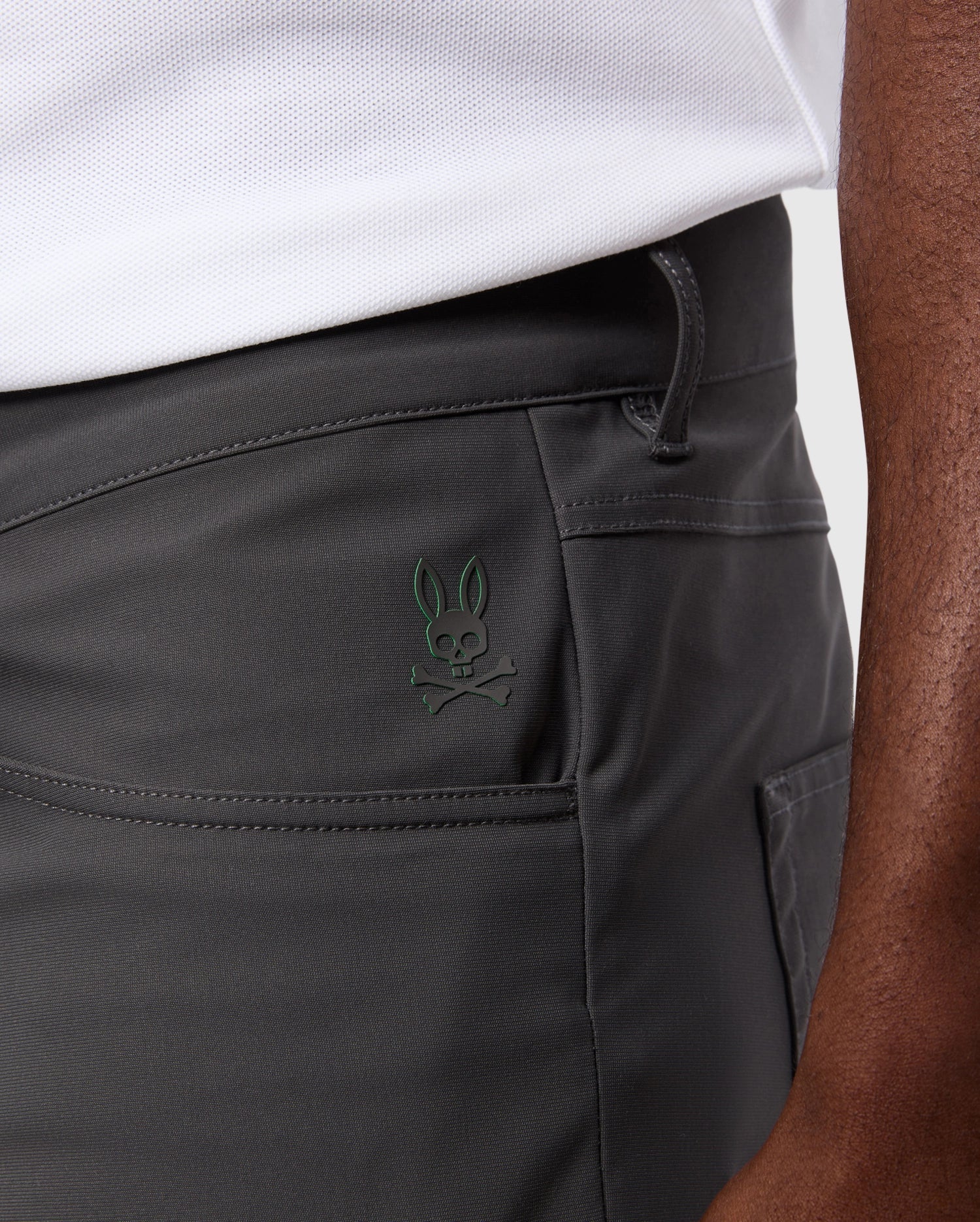 Close-up of a person wearing Psycho Bunny MENS MAVERICK 10K PANT - B6P788D200, which boasts five pockets and a small embroidered rabbit skull and crossbones logo. These dark-colored pants are designed with 4-way stretch for enhanced comfort. The person is also sporting a white shirt tucked into the pants, set against a simple, light-colored background.