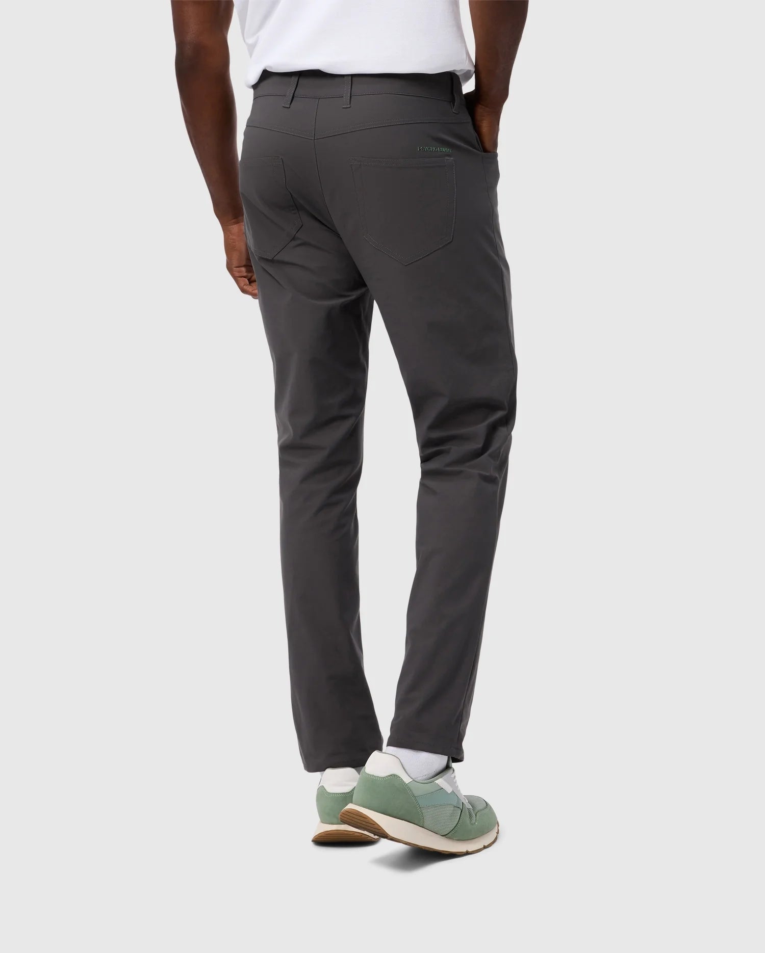 A person wearing Psycho Bunny's MENS MAVERICK 10K PANT - B6P788D200, featuring five-pocket practicality, pairs them with a white shirt and green sneakers with white soles and socks, standing back to the camera against a light gray background.