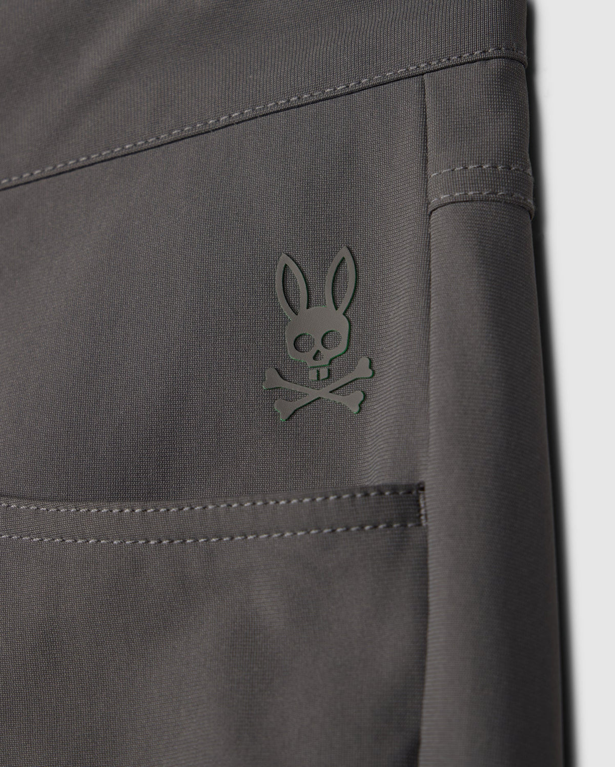 A close-up of the gray fabric from the Psycho Bunny MENS MAVERICK 10K PANT (B6P788D200) shows a stylized rabbit skull and crossbones logo embossed in dark green on the upper seam. This fabric features a 4-way stretch for added comfort.