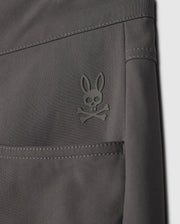 A close-up of the gray fabric from the Psycho Bunny MENS MAVERICK 10K PANT (B6P788D200) shows a stylized rabbit skull and crossbones logo embossed in dark green on the upper seam. This fabric features a 4-way stretch for added comfort.