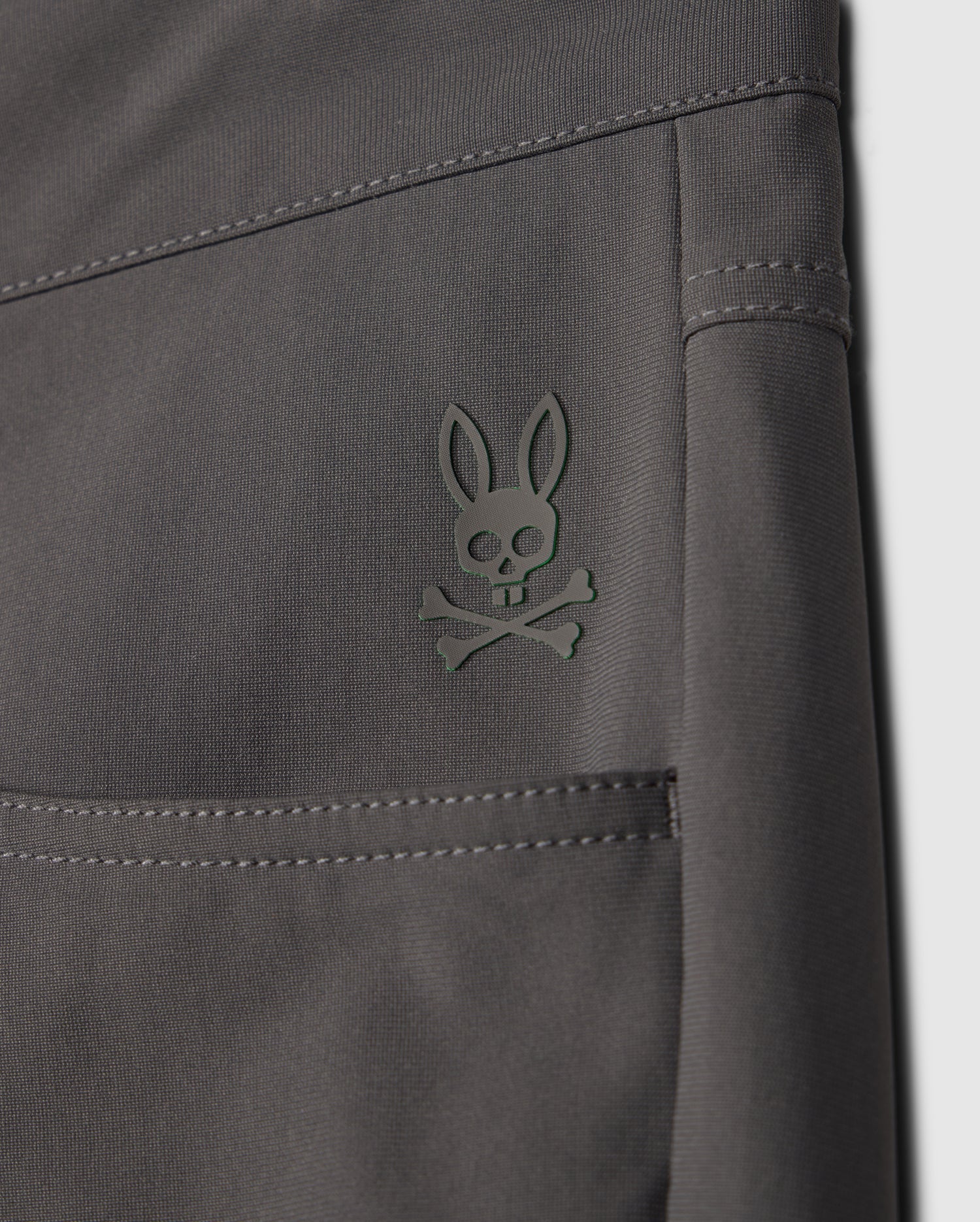 A close-up of the gray fabric from the Psycho Bunny MENS MAVERICK 10K PANT (B6P788D200) shows a stylized rabbit skull and crossbones logo embossed in dark green on the upper seam. This fabric features a 4-way stretch for added comfort.