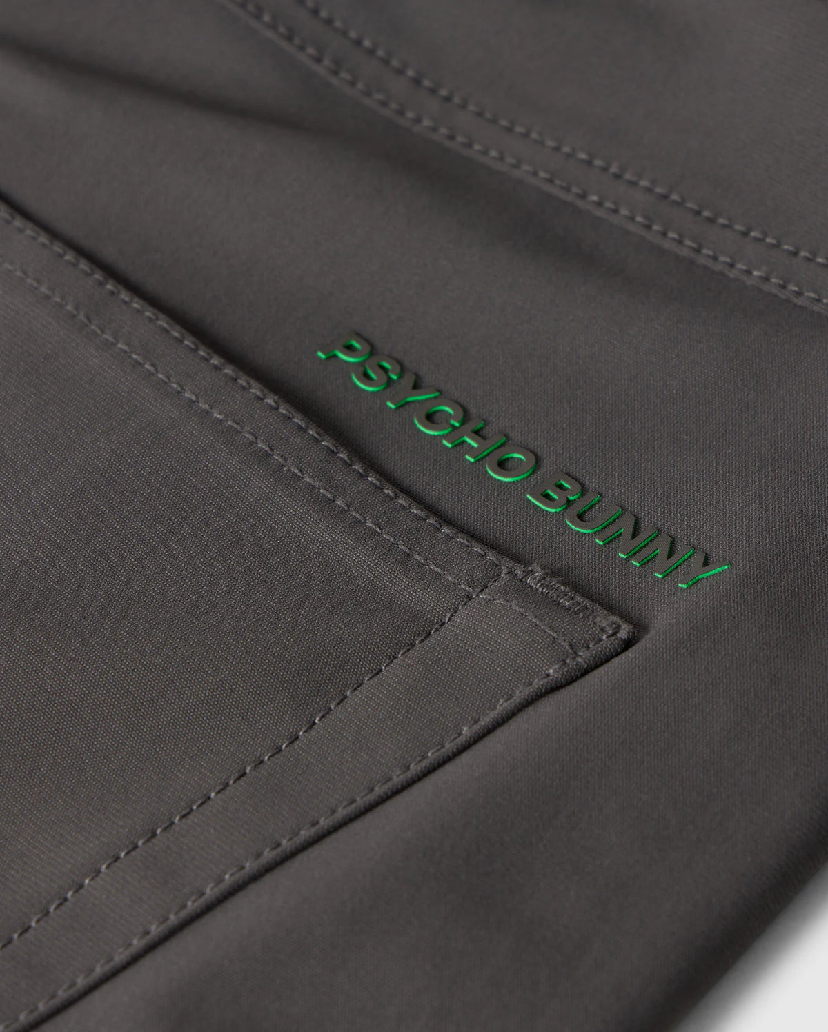 Close-up of gray fabric with visible stitching and a green "Psycho Bunny" embroidered logo on the MENS MAVERICK 10K PANT - B6P788D200, featuring five practical pockets for everyday ease.