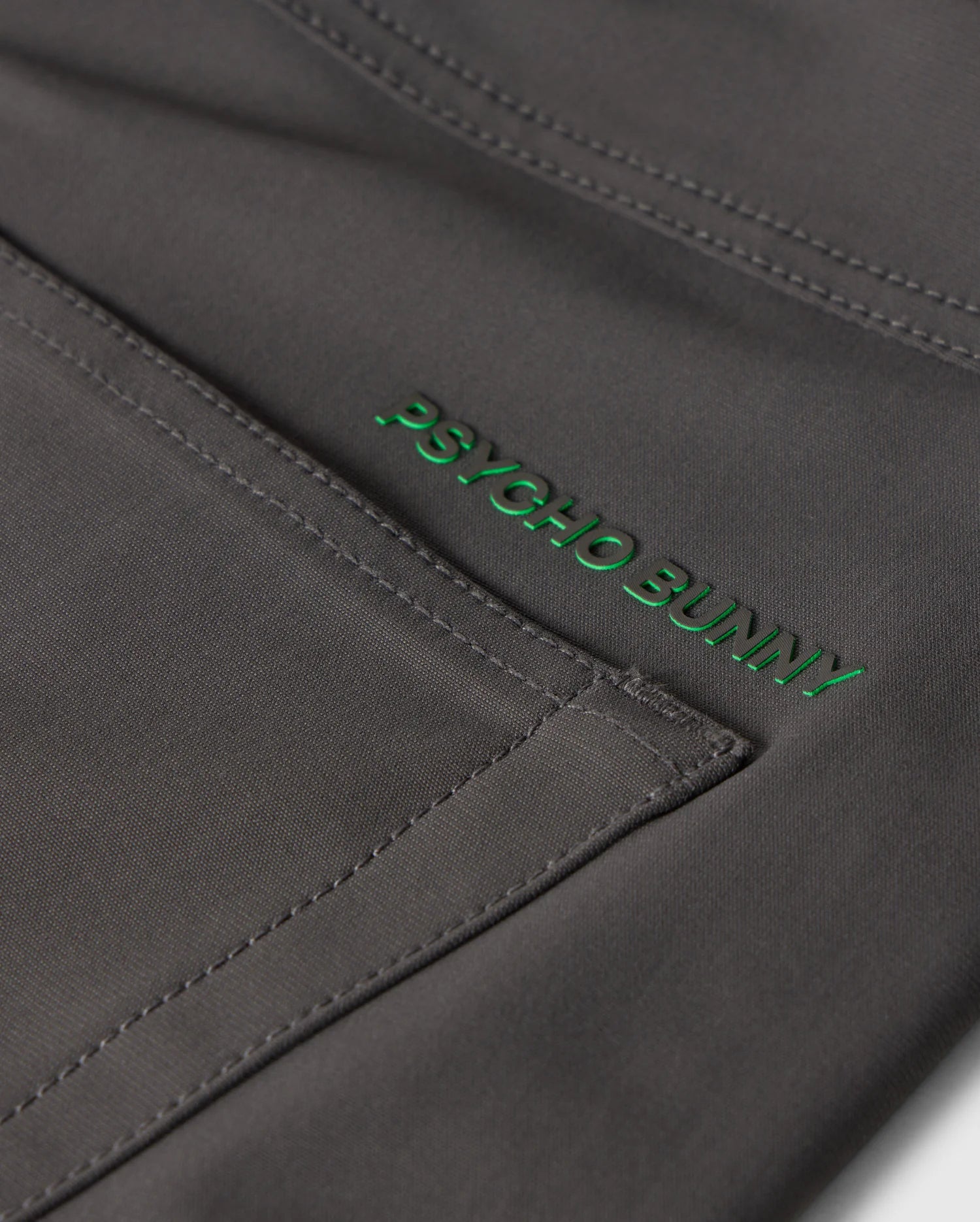 Close-up of gray fabric with visible stitching and a green "Psycho Bunny" embroidered logo on the MENS MAVERICK 10K PANT - B6P788D200, featuring five practical pockets for everyday ease.