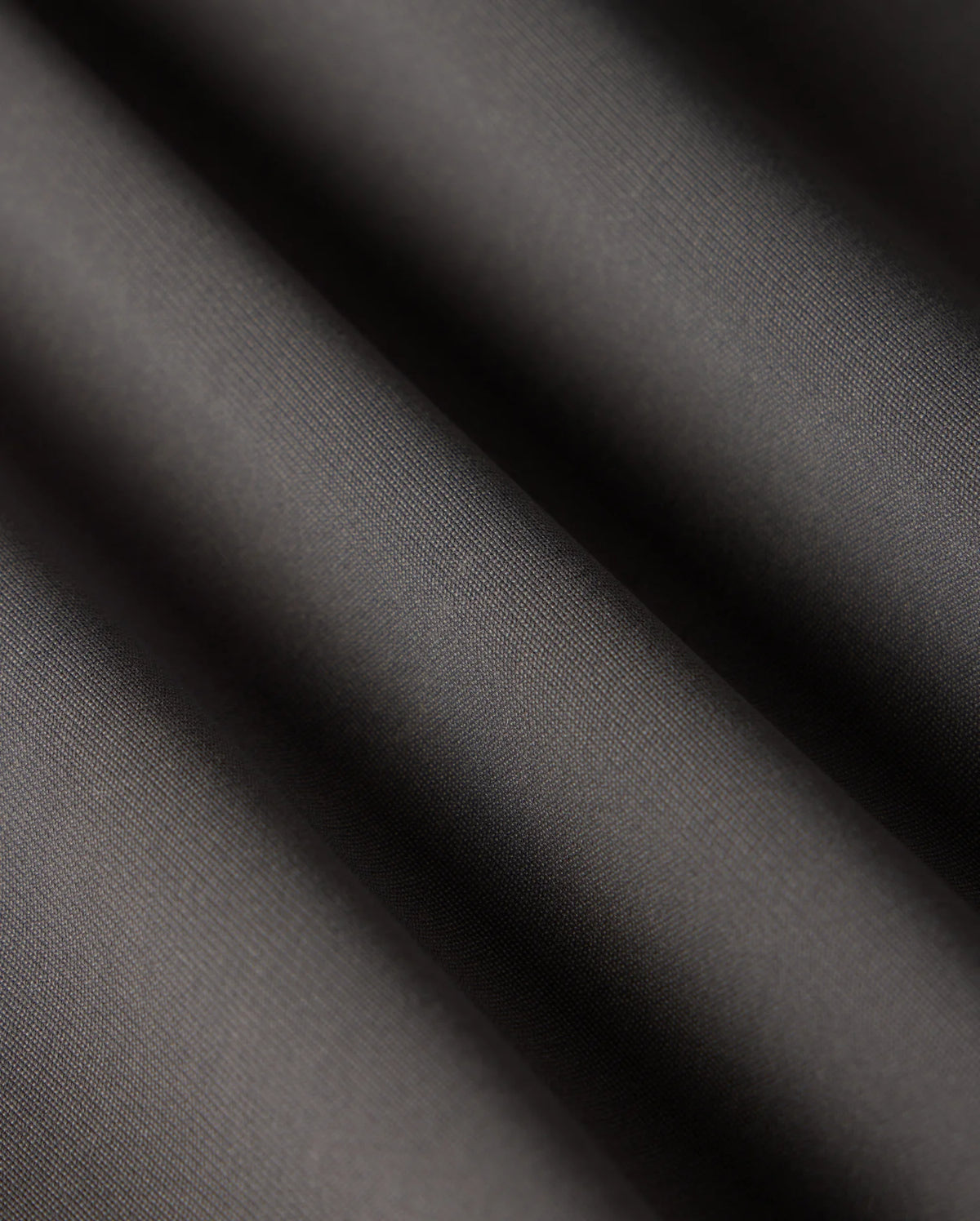 Close-up of the smooth, dark grey fabric of the Psycho Bunny MENS MAVERICK 10K PANT - B6P788D200 showcases subtle diagonal ripples and a matte finish. The folds create gentle shadows, highlighting its texture, slight sheen, and five-pocket practicality.