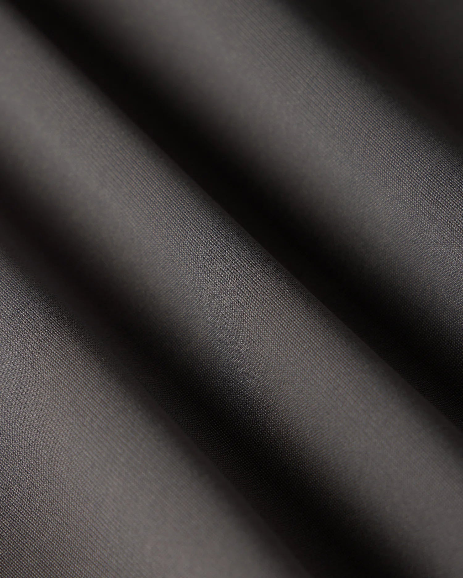 Close-up of the smooth, dark grey fabric of the Psycho Bunny MENS MAVERICK 10K PANT - B6P788D200 showcases subtle diagonal ripples and a matte finish. The folds create gentle shadows, highlighting its texture, slight sheen, and five-pocket practicality.