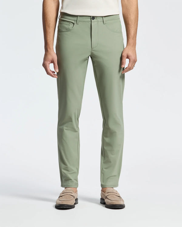 A person stands in sage green Psycho Bunny's MENS MAVERICK 10K STEP PANT - B6P788D200 with five-pocket styling, front pockets, and beige loafers. Paired with a crisp white top, the plain white background highlights its elegance and versatility.