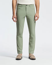 A person stands in sage green Psycho Bunny's MENS MAVERICK 10K STEP PANT - B6P788D200 with five-pocket styling, front pockets, and beige loafers. Paired with a crisp white top, the plain white background highlights its elegance and versatility.