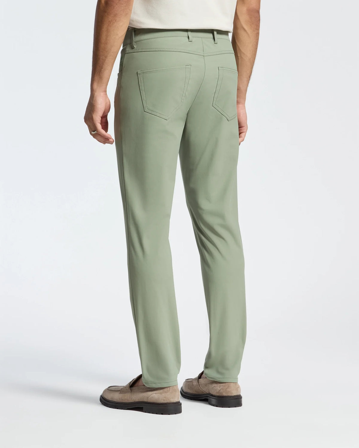 A person is shown from behind wearing Psycho Bunny's MENS MAVERICK 10K STEP PANT - B6P788D200 in sage green against a plain white background, featuring patch pockets, paired with a beige shirt and suede shoes.