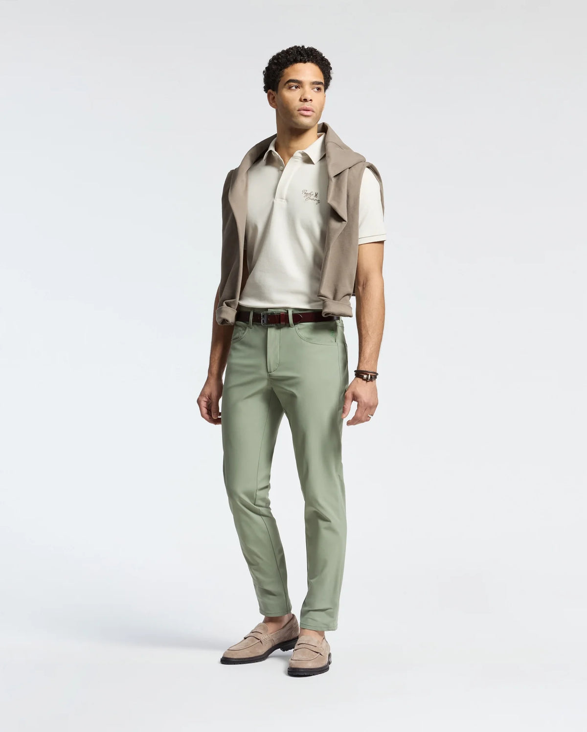 A man stands against a plain background, dressed in a white polo shirt and Psycho Bunny's MENS MAVERICK 10K STEP PANT - B6P788D200. The five-pocket styling and light green, four-way stretch fabric enhance his casual yet polished look, completed with loafers and a sweater draped over his shoulders.
