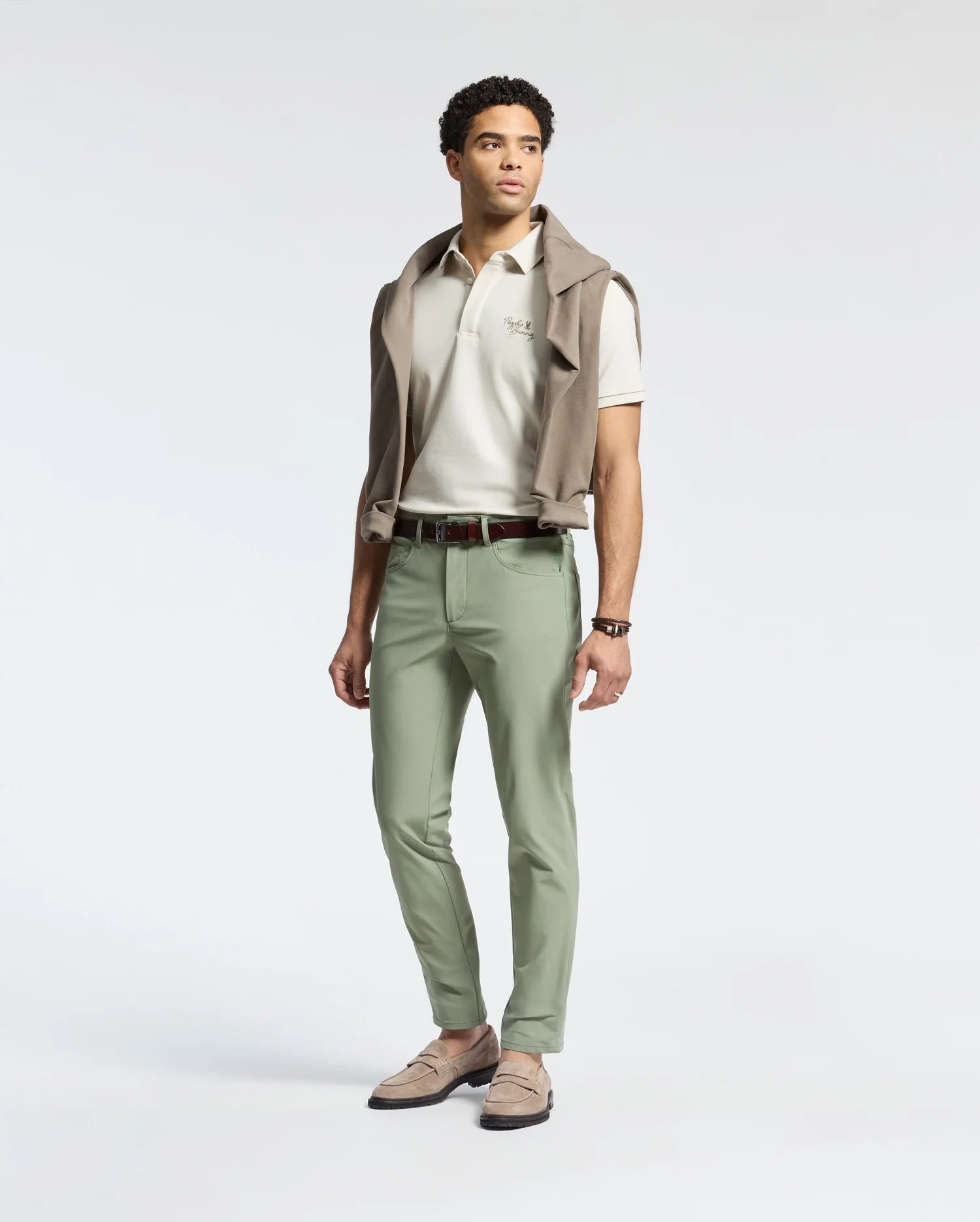 A man stands against a plain background, dressed in a white polo shirt and Psycho Bunny's MENS MAVERICK 10K STEP PANT - B6P788D200. The five-pocket styling and light green, four-way stretch fabric enhance his casual yet polished look, completed with loafers and a sweater draped over his shoulders.