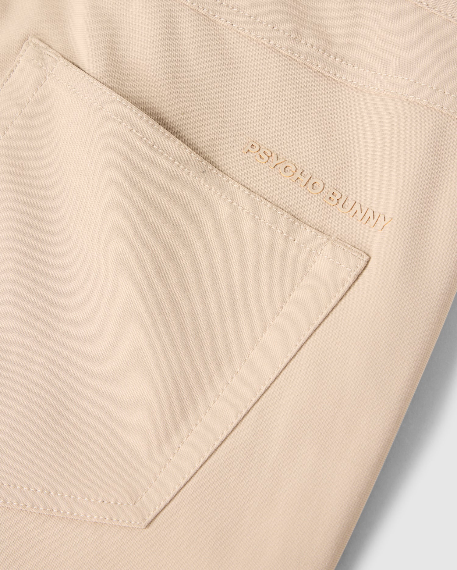 Close-up of the MENS MAVERICK 10K PANT - B6P788D200 pocket, showcasing the small embroidered "PSYCHO BUNNY" text in gold thread near the top edge. The fabric appears smooth, with clean stitching outlining the five-pocket pant's shape and offering four-way stretch for ultimate comfort.