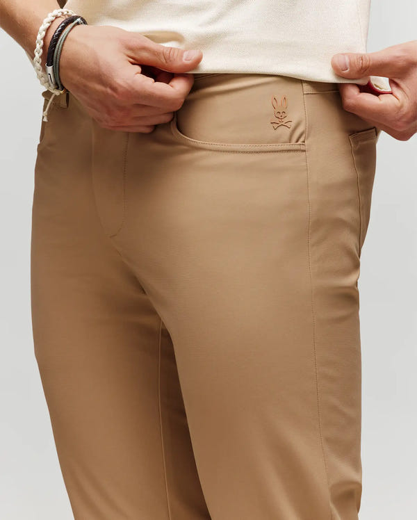 A person wearing Psycho Bunny MENS MAVERICK 10K STEP PANT (B6P788D200) in tan, featuring five-pocket styling and a small embroidered bunny logo by the front pocket, lifts their shirt slightly, bracelets on wrist.