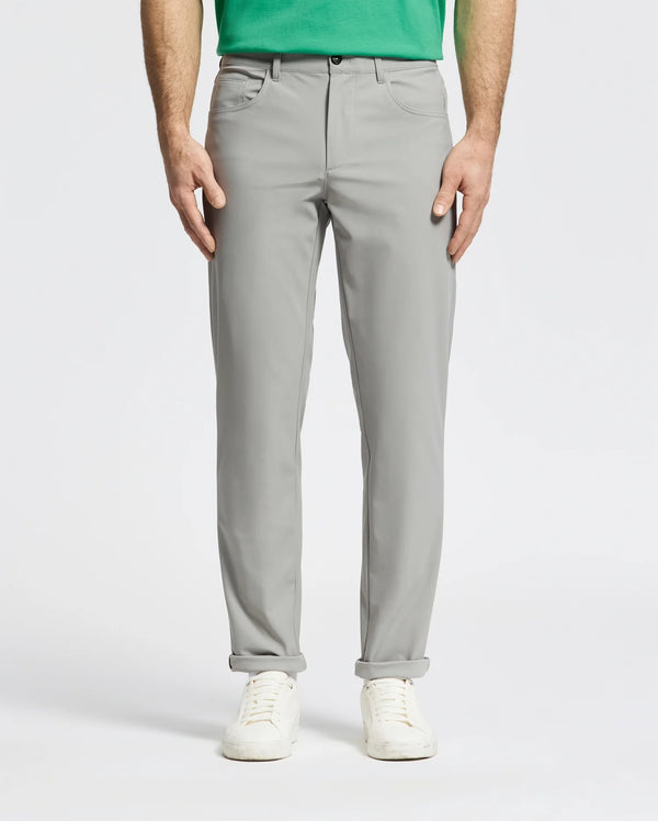 A person wearing Psycho Bunny MENS MAVERICK 10K STEP PANT - B6P788D200 with five-pocket styling, a green shirt, and white sneakers stands hands-in-pockets against a plain light background; only the lower half is visible.