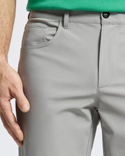 A close-up shows the lower torso and relaxed left hand of a person wearing Psycho Bunny MENS MAVERICK 10K STEP PANT - B6P788D200 in light gray, highlighting the five-pocket design near the side pocket. Only part of a green shirt is visible.