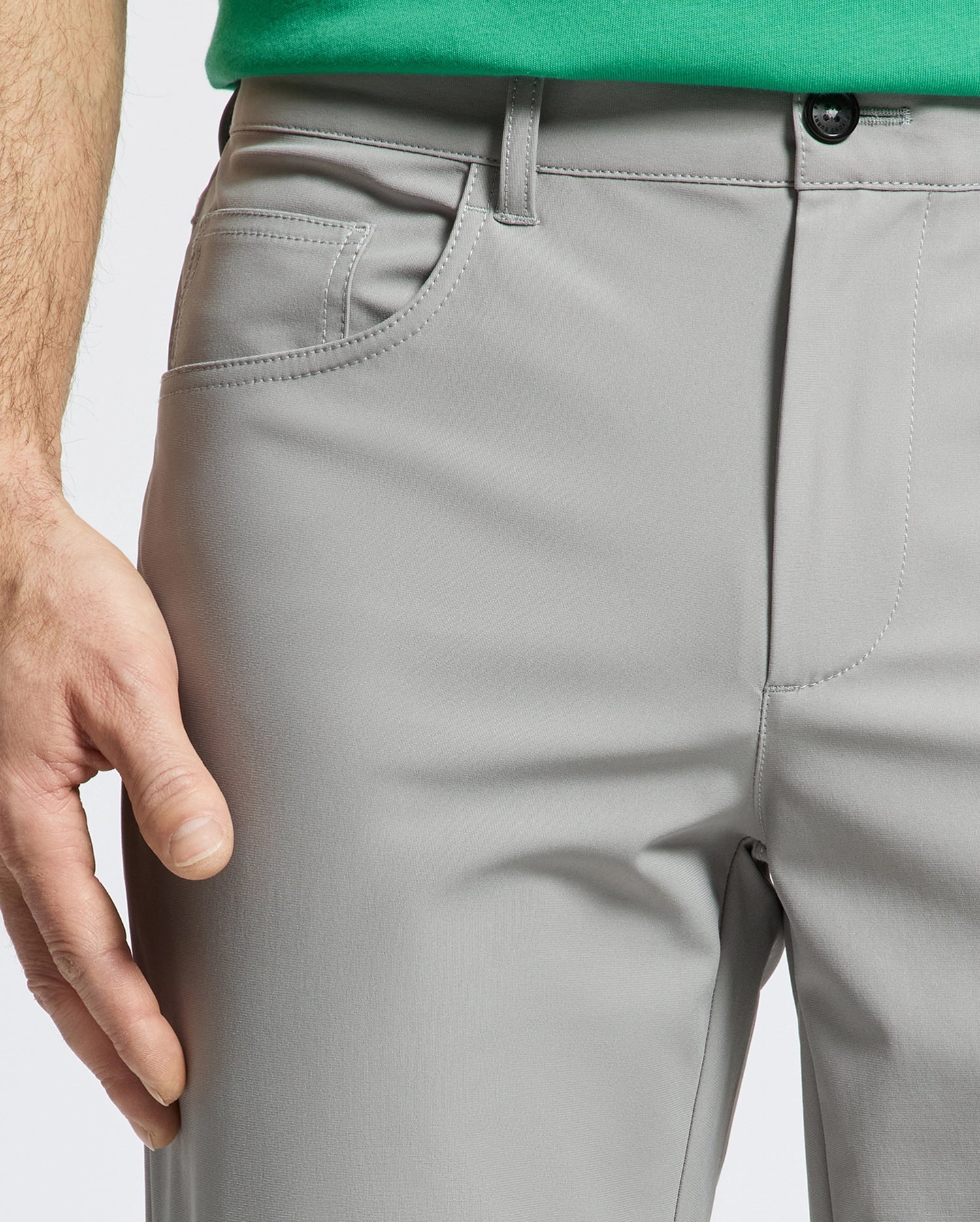 A close-up shows the lower torso and relaxed left hand of a person wearing Psycho Bunny MENS MAVERICK 10K STEP PANT - B6P788D200 in light gray, highlighting the five-pocket design near the side pocket. Only part of a green shirt is visible.