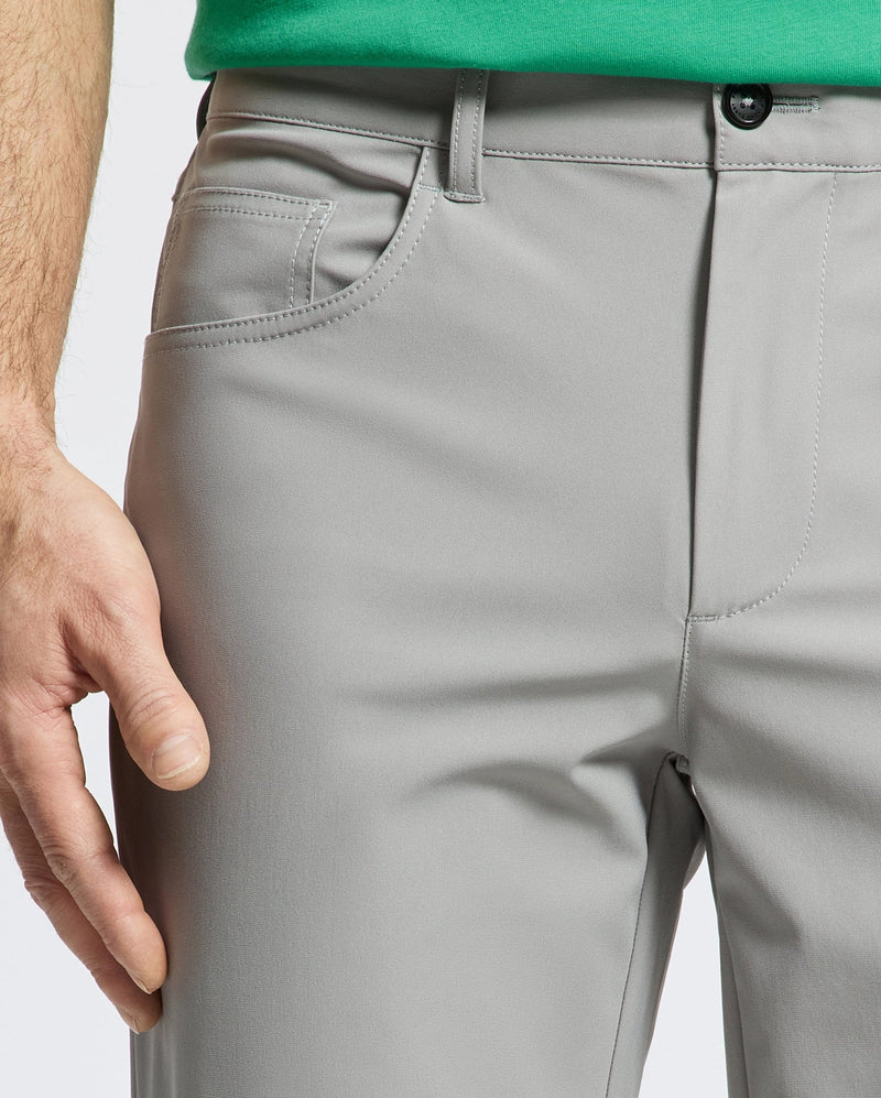 A close-up shows the lower torso and relaxed left hand of a person wearing Psycho Bunny MENS MAVERICK 10K STEP PANT - B6P788D200 in light gray, highlighting the five-pocket design near the side pocket. Only part of a green shirt is visible.