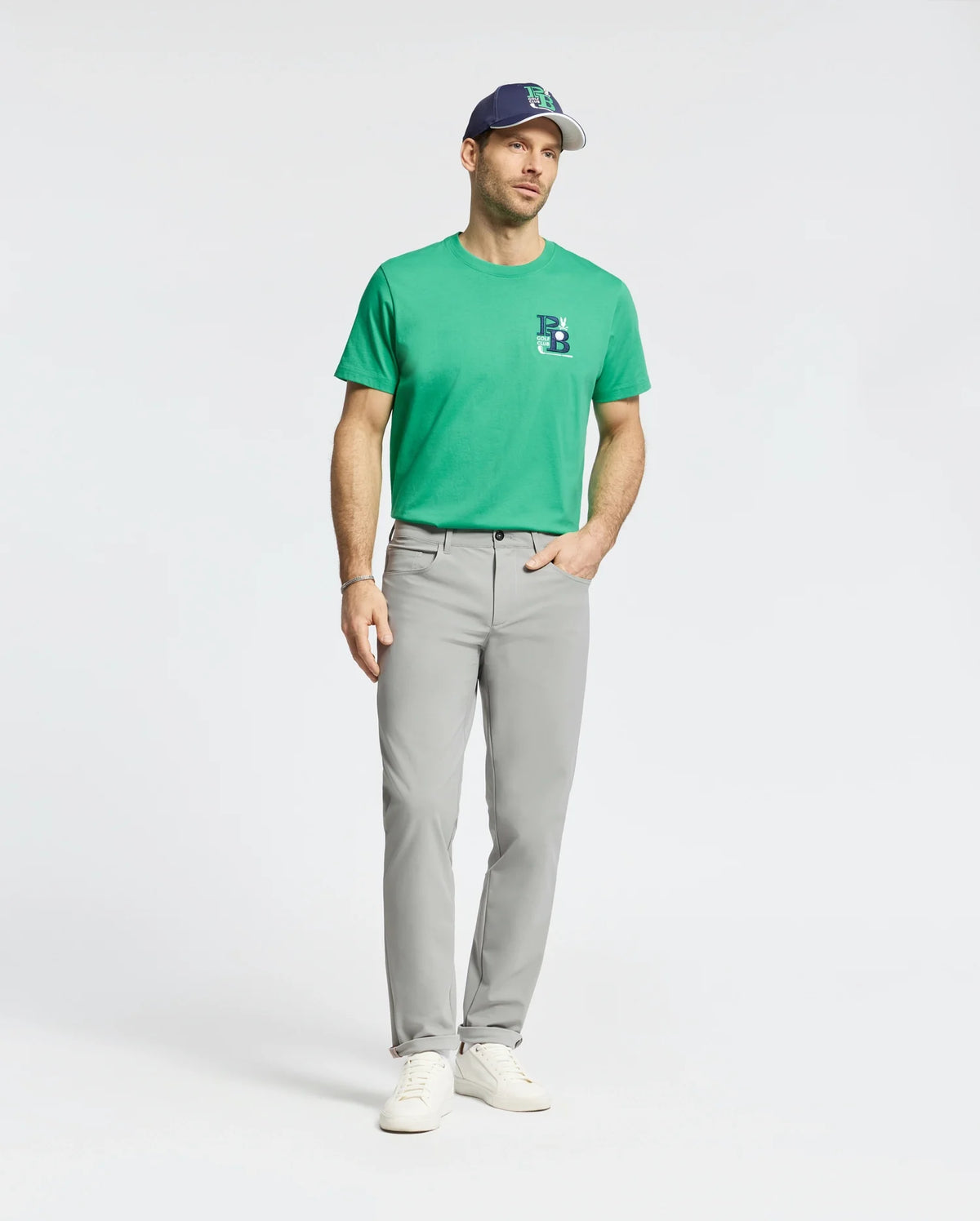 A man wears a green Psycho Bunny tee, white sneakers, a blue and white cap, and the MENS MAVERICK 10K STEP PANT - B6P788D200 featuring five-pocket styling and four-way stretch, standing against a plain white background.