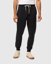 A person is wearing the Psycho Bunny MENS JAMES PREMIUM SWEATPANT (B6P791D200) in black, made from soft cotton-blend fabric, tied with a beige drawstring, paired with a white shirt and beige sneakers that have white laces. The person is standing against a plain light grey background.