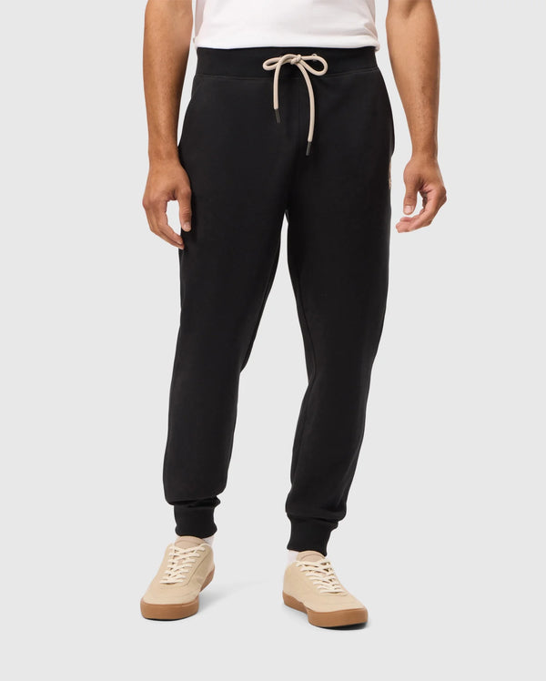 A person is wearing the Psycho Bunny MENS JAMES PREMIUM SWEATPANT (B6P791D200) in black, made from soft cotton-blend fabric, tied with a beige drawstring, paired with a white shirt and beige sneakers that have white laces. The person is standing against a plain light grey background.