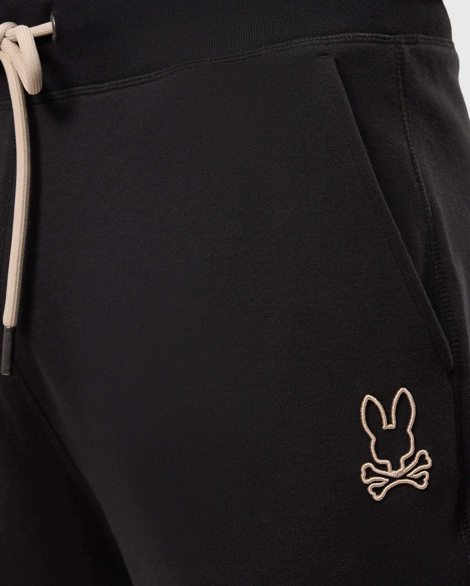Close-up of the upper section of a pair of MENS JAMES PREMIUM SWEATPANT - B6P791D200 by Psycho Bunny, showcasing black sweatpants with an elastic waistband and beige drawstrings. Crafted from a soft cotton-blend for ultimate comfort, these premium bottoms feature an embroidered beige logo of a bunny head with crossbones just below the front right pocket.