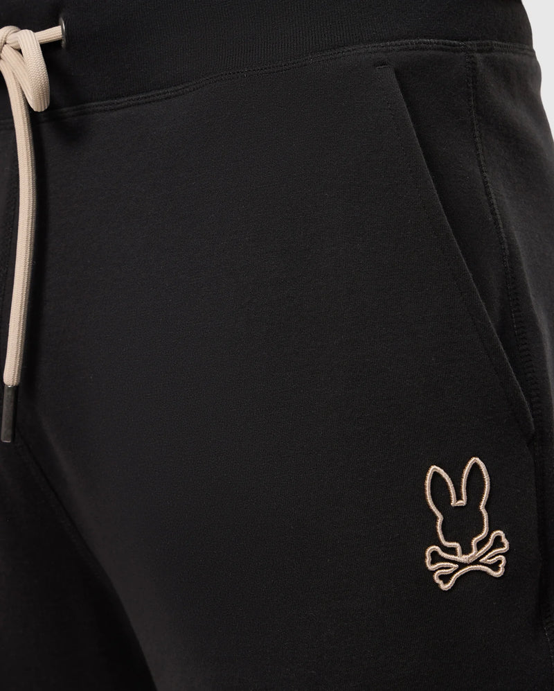 Close-up of the upper section of a pair of MENS JAMES PREMIUM SWEATPANT - B6P791D200 by Psycho Bunny, showcasing black sweatpants with an elastic waistband and beige drawstrings. Crafted from a soft cotton-blend for ultimate comfort, these premium bottoms feature an embroidered beige logo of a bunny head with crossbones just below the front right pocket.