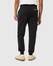 A person is seen from the back, wearing a white t-shirt and dark jogger pants from Psycho Bunny's MENS JAMES PREMIUM SWEATPANT - B6P791D200 collection, with "PSYCHO BUNNY" written on the right pocket. These premium bottoms, crafted from a soft cotton-blend, promise ultimate comfort. They are also wearing off-white sneakers with brown soles. The background is plain and light gray.