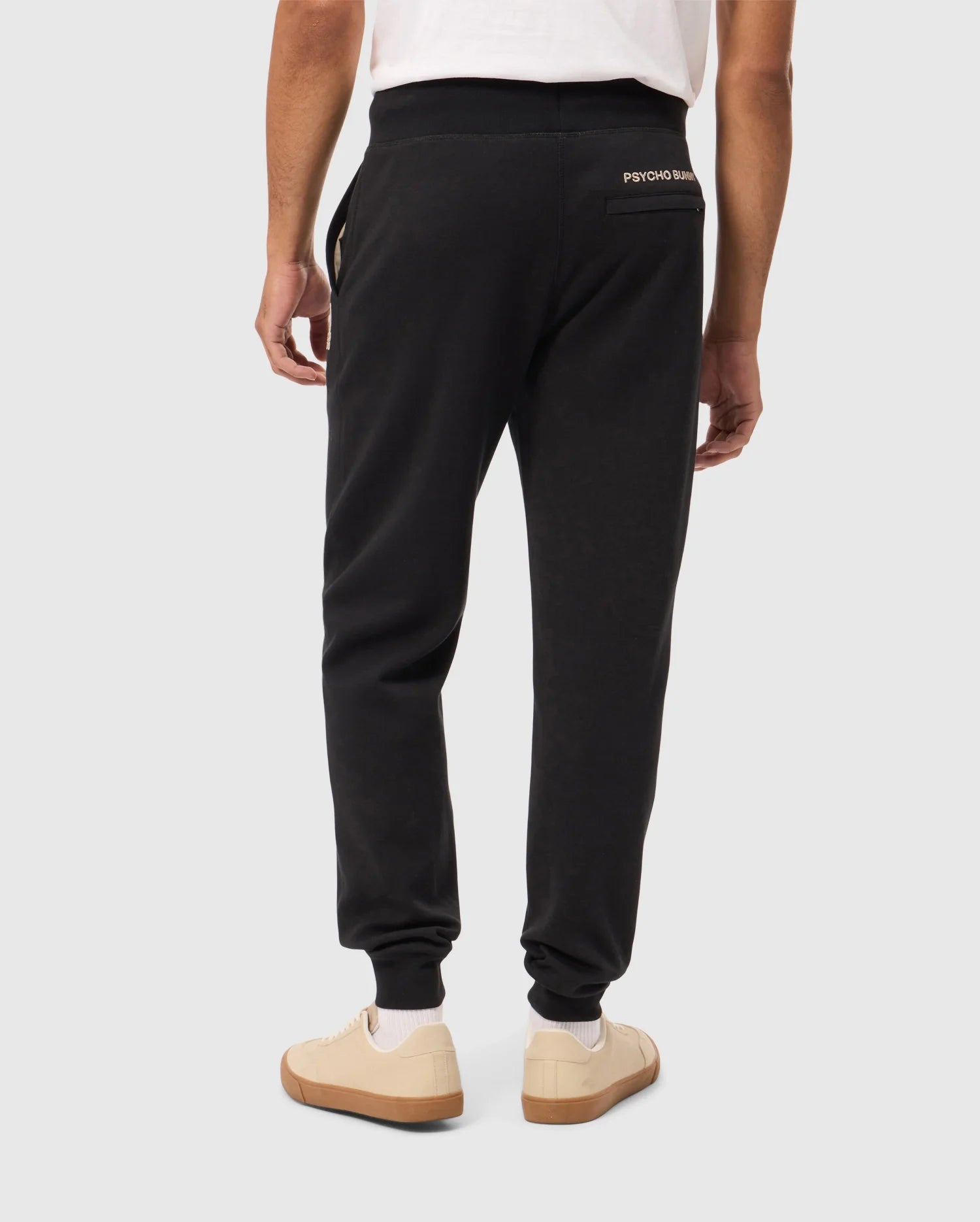 A person is seen from the back, wearing a white t-shirt and dark jogger pants from Psycho Bunny's MENS JAMES PREMIUM SWEATPANT - B6P791D200 collection, with "PSYCHO BUNNY" written on the right pocket. These premium bottoms, crafted from a soft cotton-blend, promise ultimate comfort. They are also wearing off-white sneakers with brown soles. The background is plain and light gray.
