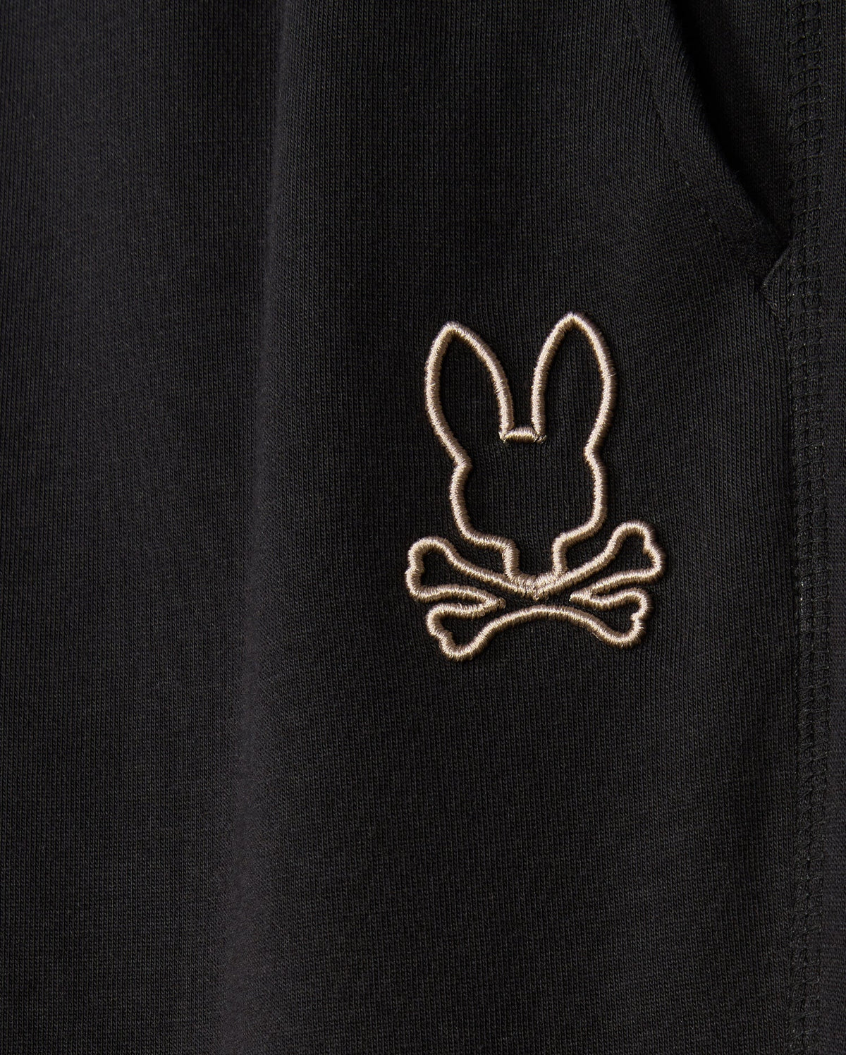 Close-up image of black, soft cotton-blend fabric on the MENS JAMES PREMIUM SWEATPANT - B6P791D200 by Psycho Bunny, featuring an embroidered design of a bunny head above two crossed bones, stitched in light-colored thread.