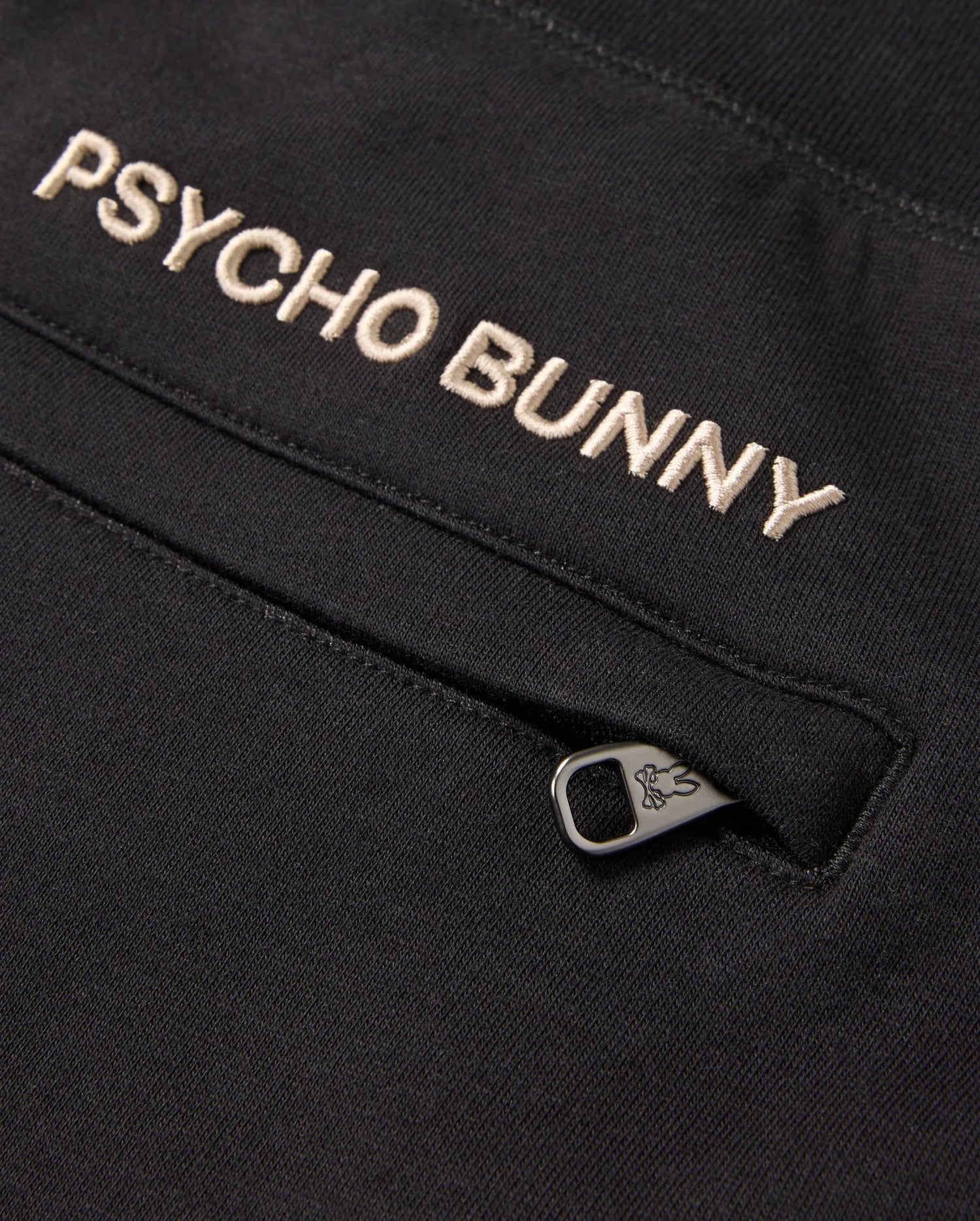 Close-up of a black fabric with "Psycho Bunny" embroidered in white thread. Below the text is a zipper with a small bunny icon on the metal pull tab. Made from a soft cotton-blend for ultimate comfort, the fabric is part of the MENS JAMES PREMIUM SWEATPANT - B6P791D200 by Psycho Bunny.