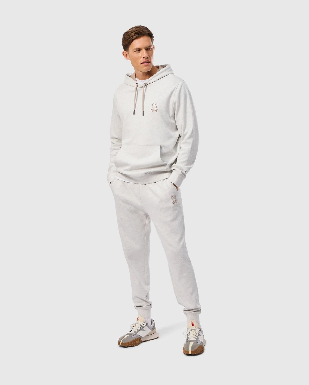 A person dons the MENS JAMES PREMIUM SWEATPANT in light gray cotton-blend by Psycho Bunny. They stand with hands in pockets, sporting an outfit that includes a subtle embroidered design. Complementing this look are sneakers with gray and beige accents on a plain background, highlighting ultimate comfort.
