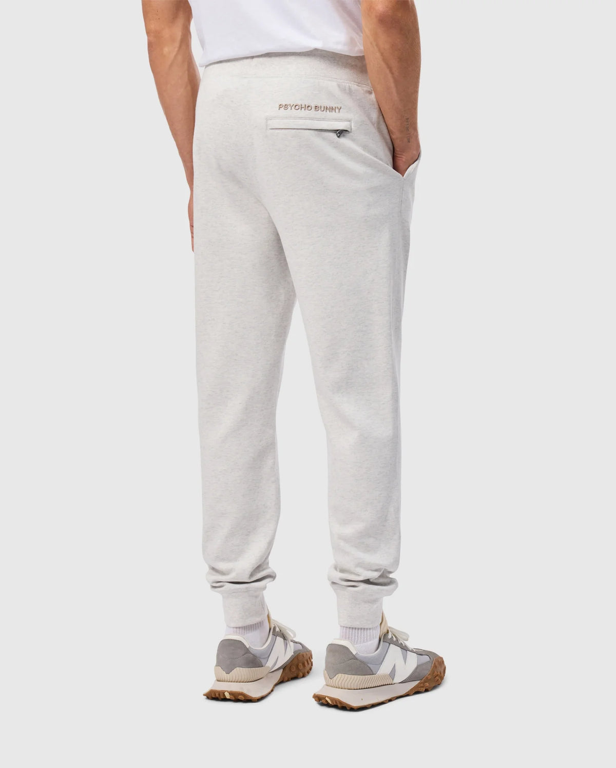 A person dressed in a white T-shirt is sporting the MENS JAMES PREMIUM SWEATPANT - B6P791D200 by Psycho Bunny, made from a comfortable cotton blend in light gray. With hands tucked into the pockets, the sweatpants highlight a subtle "Psycho Bunny" logo on the back and are combined with gray and white sneakers featuring brown soles against a simple background.