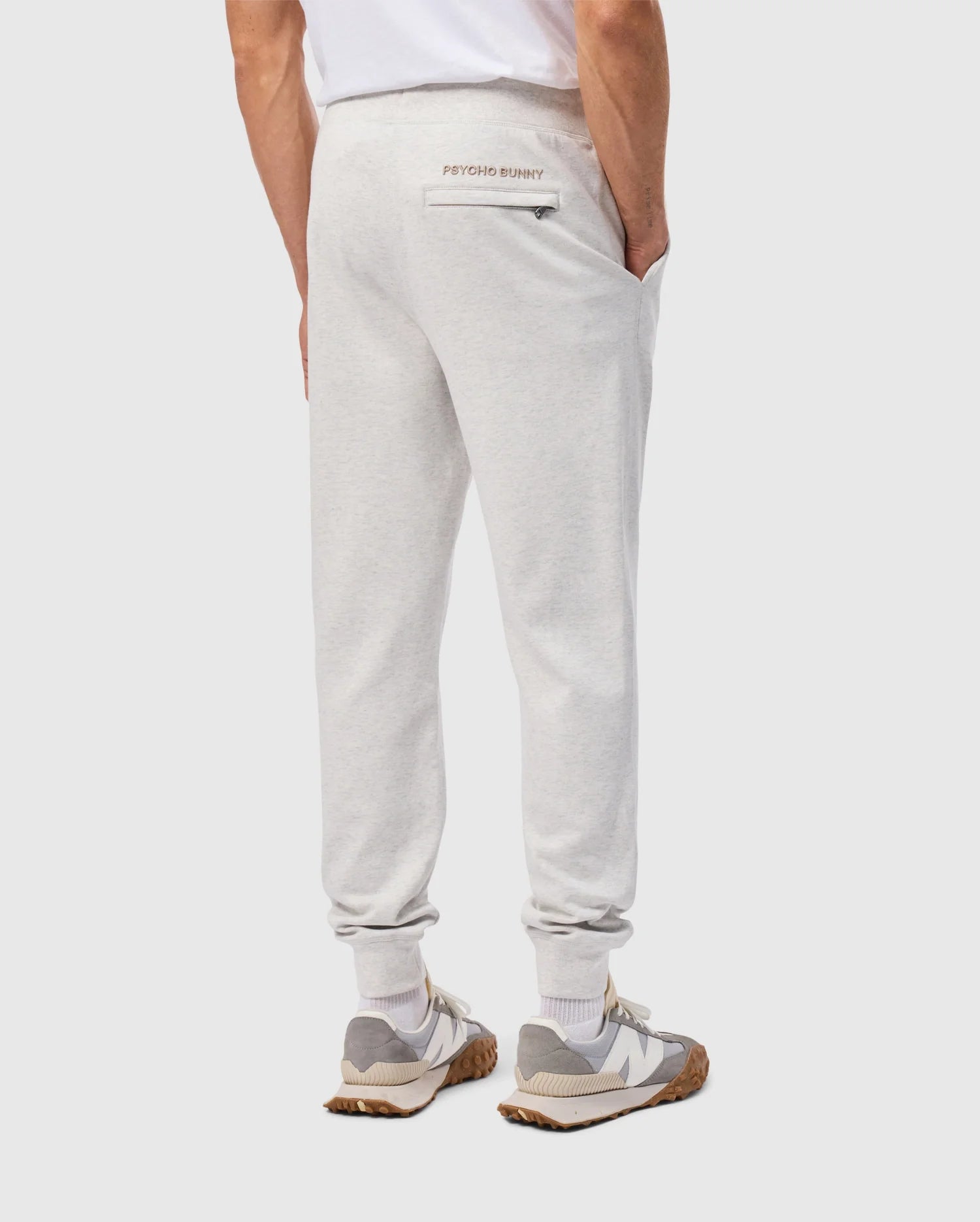 A person dressed in a white T-shirt is sporting the MENS JAMES PREMIUM SWEATPANT - B6P791D200 by Psycho Bunny, made from a comfortable cotton blend in light gray. With hands tucked into the pockets, the sweatpants highlight a subtle "Psycho Bunny" logo on the back and are combined with gray and white sneakers featuring brown soles against a simple background.