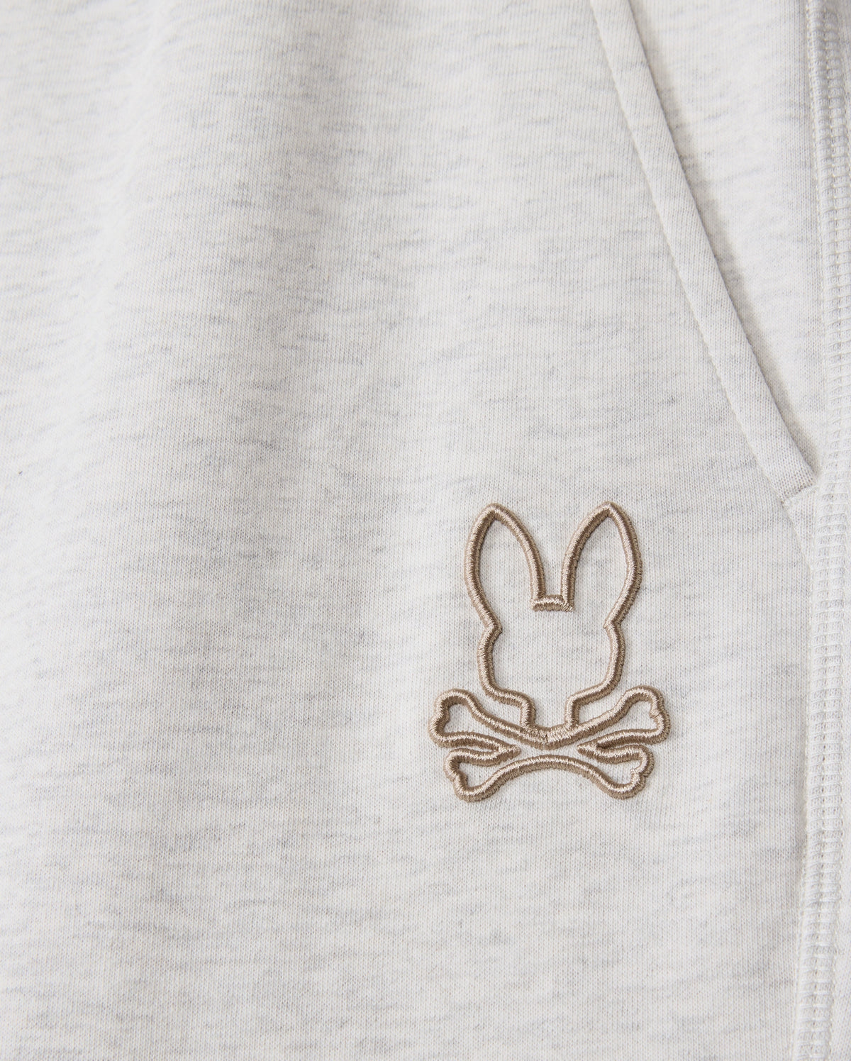 Zoom in on the soft, heathered cotton-blend fabric of the MENS JAMES PREMIUM SWEATPANT - B6P791D200 from Psycho Bunny, featuring a stitched emblem. The bunny head with elongated ears above crossed bones resembles a pirate symbol in light brown thread, offering perfect comfort for any sweatpant design.