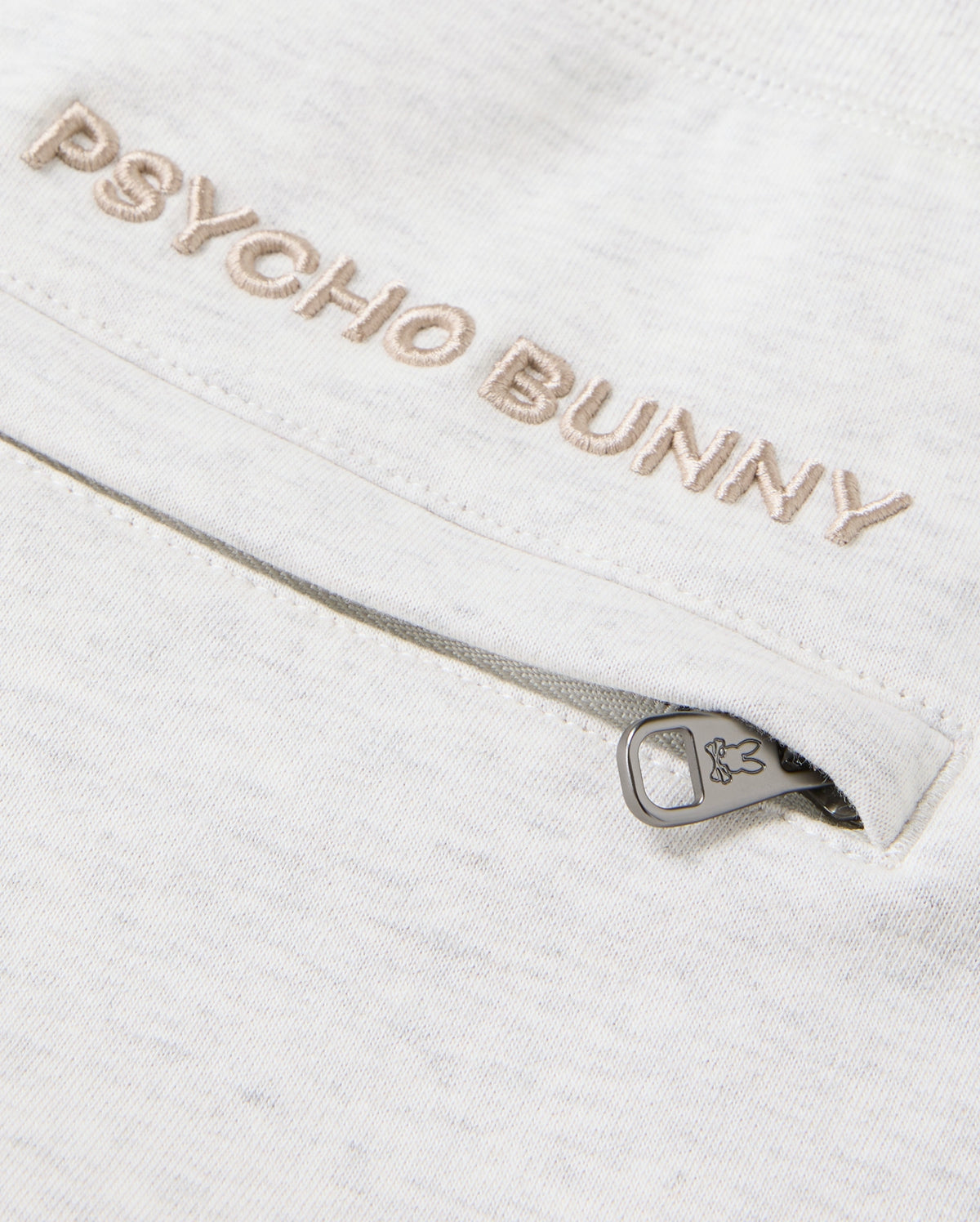 Close-up of the MENS JAMES PREMIUM SWEATPANT in a light-colored cotton-blend, featuring "Psycho Bunny" embroidered above a zipped pocket. The zipper pull is adorned with a subtle bunny logo, enhancing the comfort of this sleek design by Psycho Bunny.