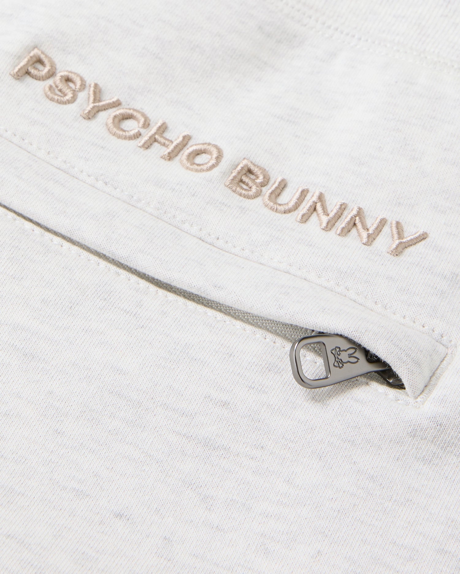 Close-up of the MENS JAMES PREMIUM SWEATPANT in a light-colored cotton-blend, featuring "Psycho Bunny" embroidered above a zipped pocket. The zipper pull is adorned with a subtle bunny logo, enhancing the comfort of this sleek design by Psycho Bunny.