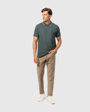 A person wearing a dark green polo shirt made from Cotton-Blend Twill, Psycho Bunny MENS HAYS TWILL PANT in beige, and white sneakers poses against a plain white background. The shirt, known for offering Ultimate Comfort with its small logo on the chest, has one hand in their pocket.