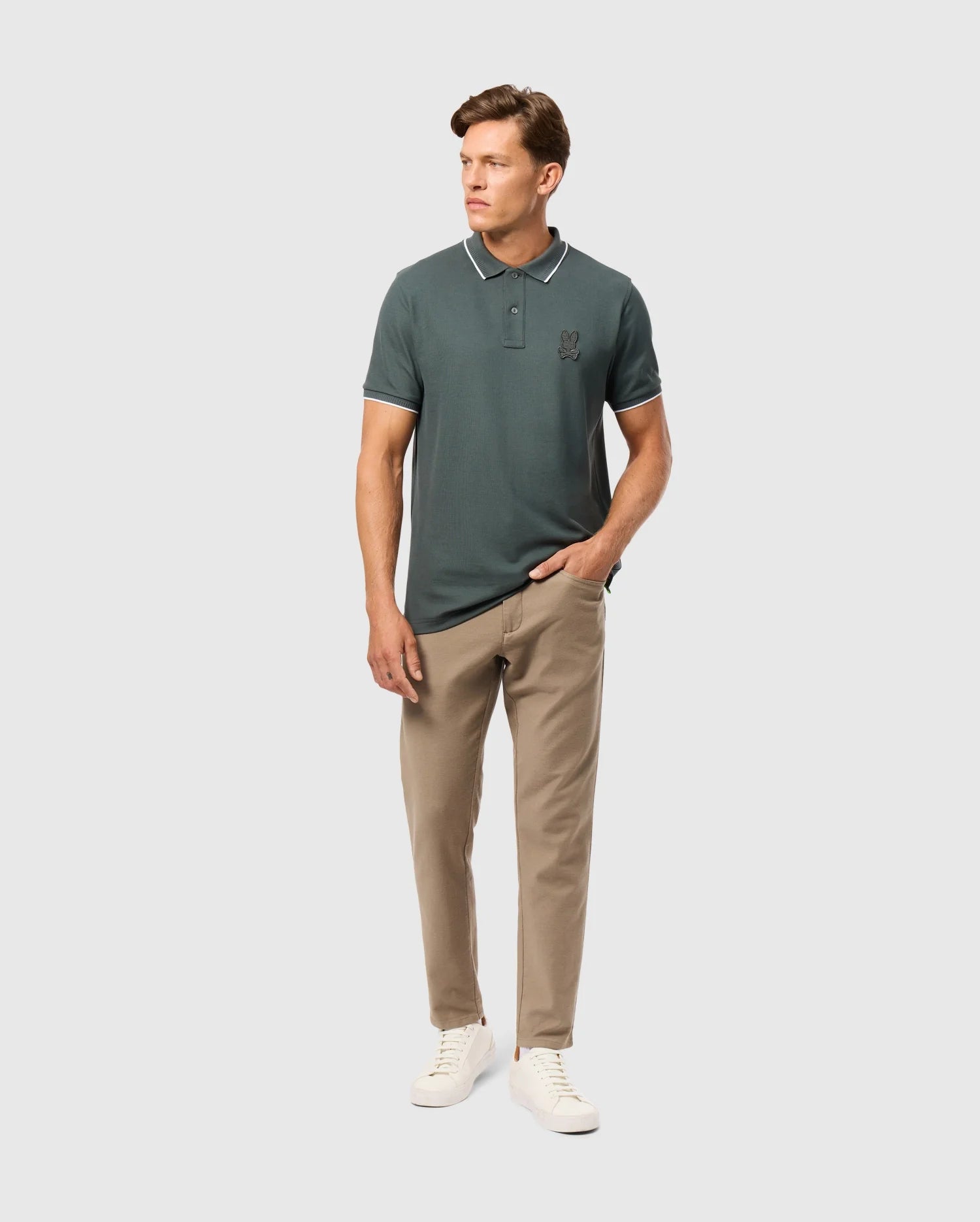 A person wearing a dark green polo shirt made from Cotton-Blend Twill, Psycho Bunny MENS HAYS TWILL PANT in beige, and white sneakers poses against a plain white background. The shirt, known for offering Ultimate Comfort with its small logo on the chest, has one hand in their pocket.