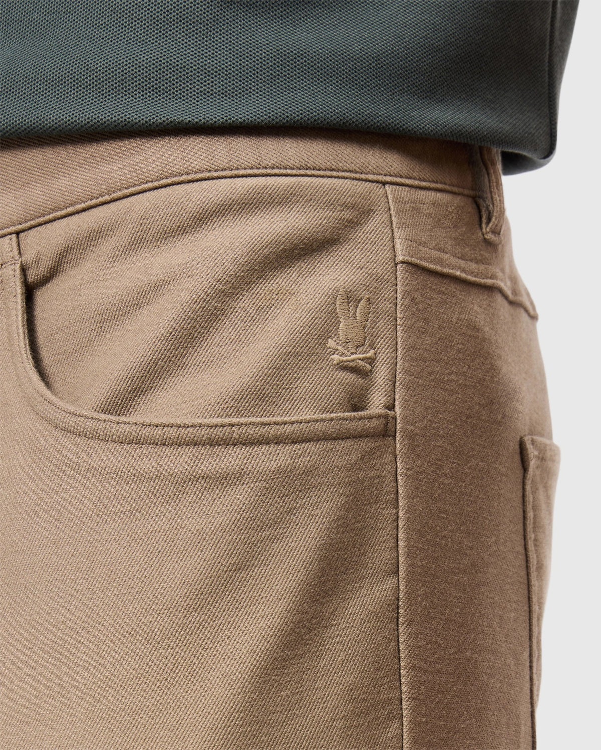 Close-up of the MENS HAYS TWILL PANT by Psycho Bunny, crafted from a cotton-blend twill in a tan color with a pocket featuring an embroidered bunny logo. The pants are designed for ultimate comfort and are worn with a dark-colored shirt tucked in, highlighting the garment's intricate stitching details.
