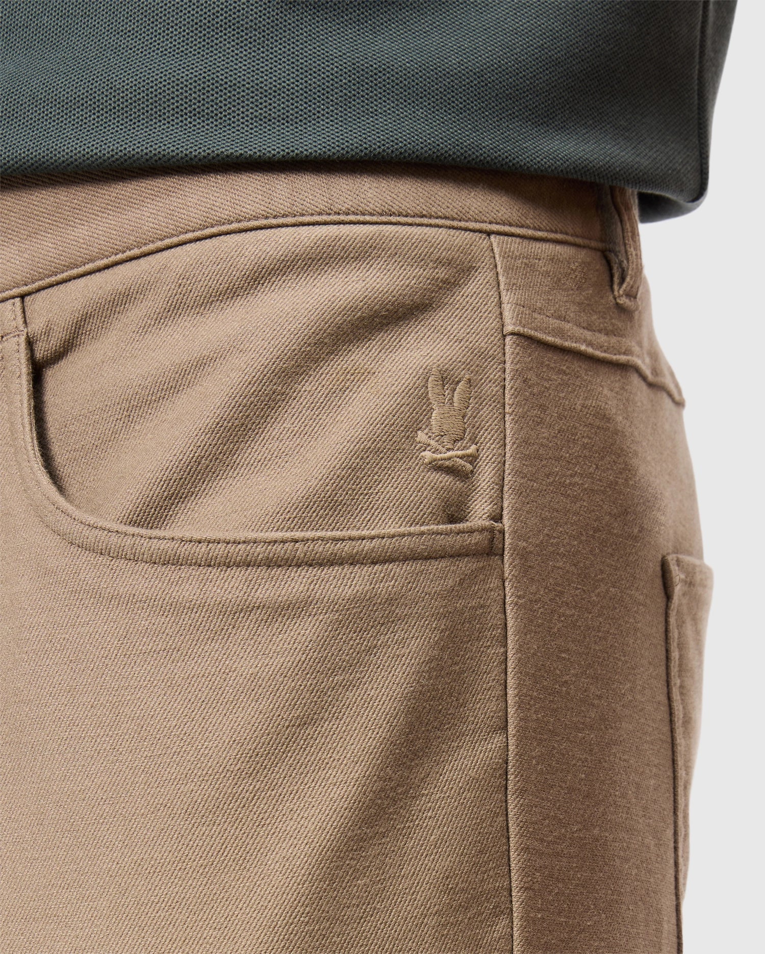 Close-up of the MENS HAYS TWILL PANT by Psycho Bunny, crafted from a cotton-blend twill in a tan color with a pocket featuring an embroidered bunny logo. The pants are designed for ultimate comfort and are worn with a dark-colored shirt tucked in, highlighting the garment's intricate stitching details.