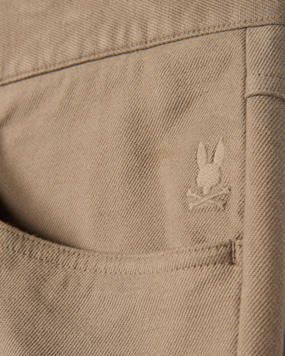 Close-up of the tan twill fabric from Psycho Bunny's MENS HAYS TWILL PANT - B6P894D200, featuring a back pocket adorned with an embroidered logo of a rabbit's head above crossed bones. The detailed stitching enhances the texture, ensuring ultimate comfort.