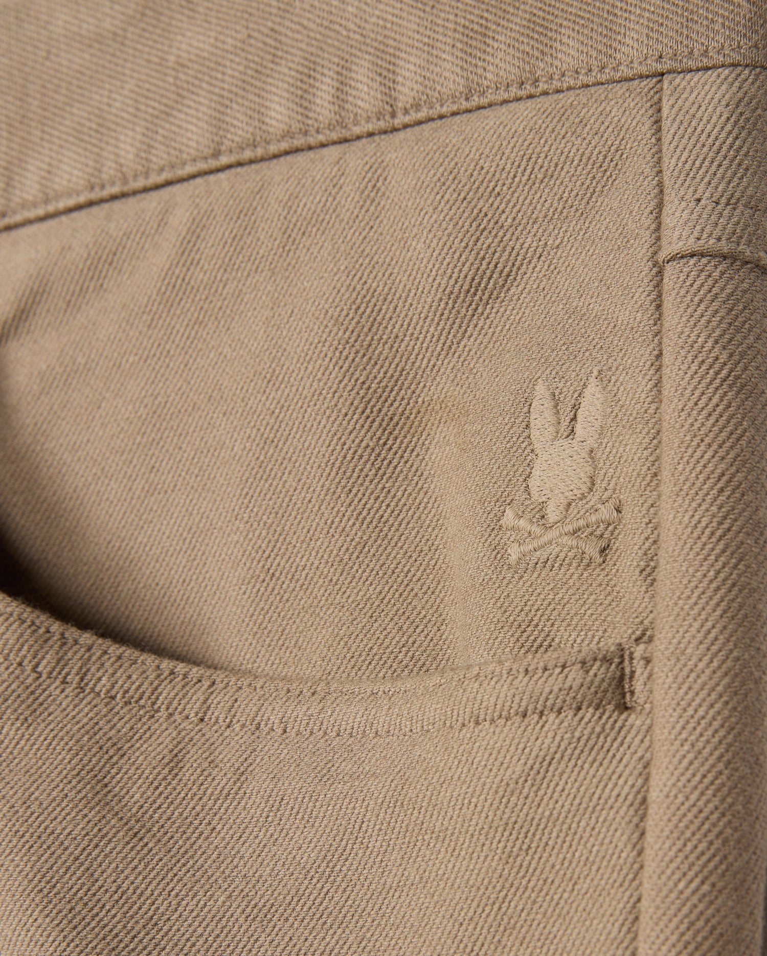 Close-up of the tan twill fabric from Psycho Bunny's MENS HAYS TWILL PANT - B6P894D200, featuring a back pocket adorned with an embroidered logo of a rabbit's head above crossed bones. The detailed stitching enhances the texture, ensuring ultimate comfort.