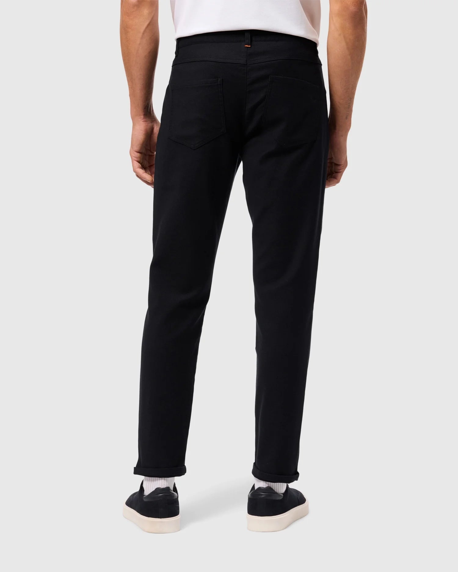 Rear view of a person showcasing ultimate comfort in Psycho Bunny's MENS HAYS TWILL PANT - B6P894D200, paired with sleek black sneakers featuring white soles, and a white shirt. The ensemble stands out against a light grey background.