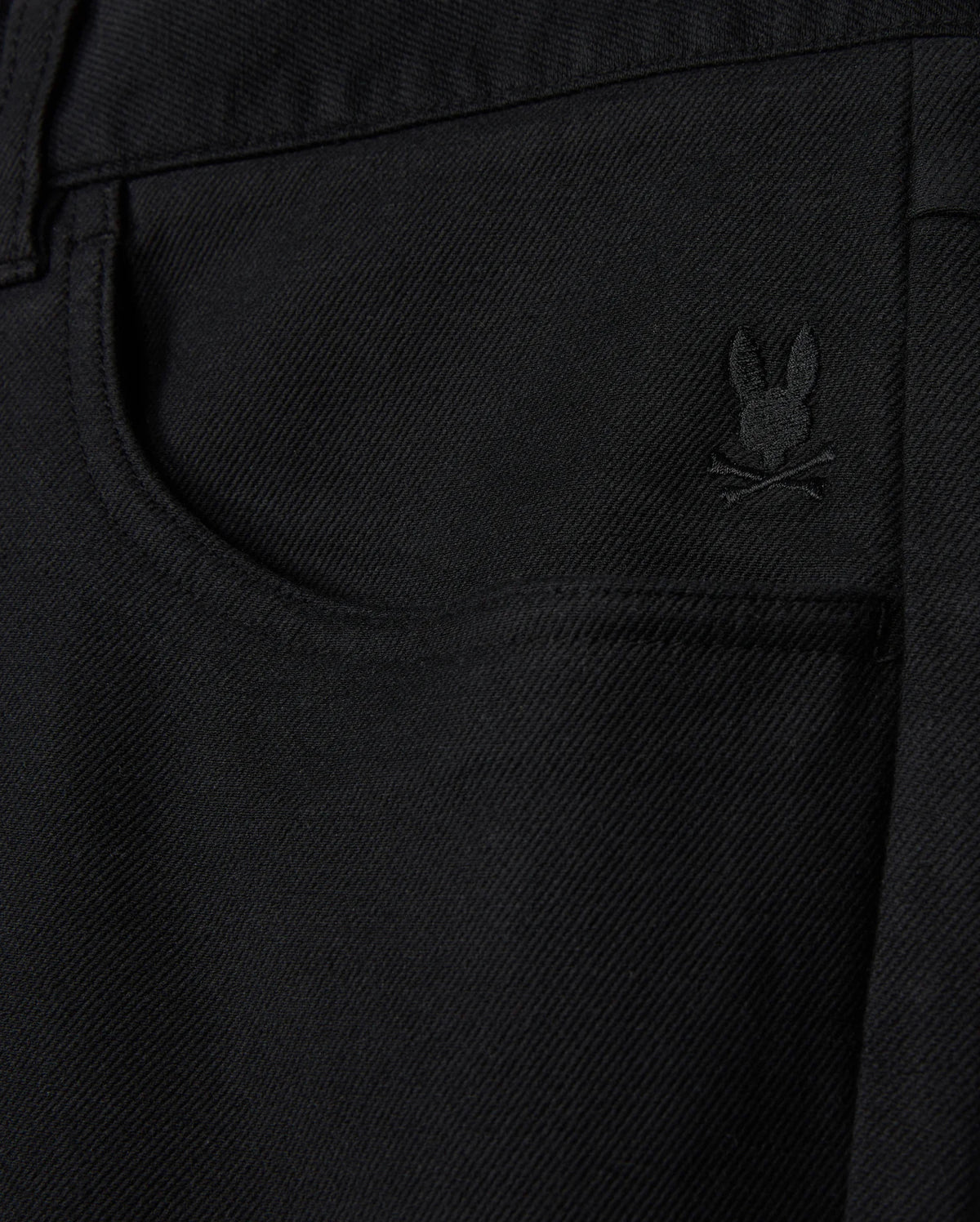The MENS HAYS TWILL PANT by Psycho Bunny showcases a close-up of black denim fabric, highlighted by a small embroidered logo of a bunny above crossed bones located near the front pocket. This cotton-blend twill design provides ultimate comfort and flexibility with its 4-way stretch, delivering both style and practicality.