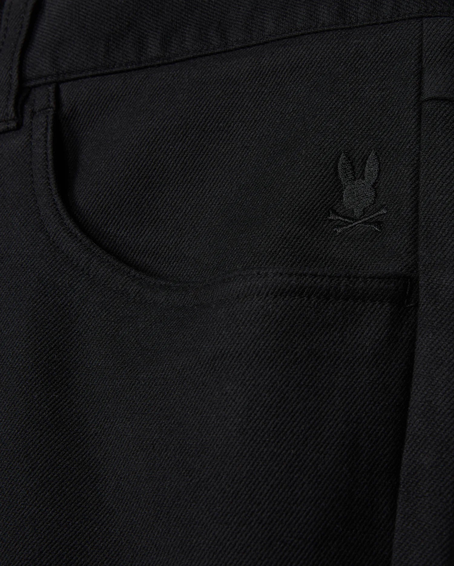 The MENS HAYS TWILL PANT by Psycho Bunny showcases a close-up of black denim fabric, highlighted by a small embroidered logo of a bunny above crossed bones located near the front pocket. This cotton-blend twill design provides ultimate comfort and flexibility with its 4-way stretch, delivering both style and practicality.