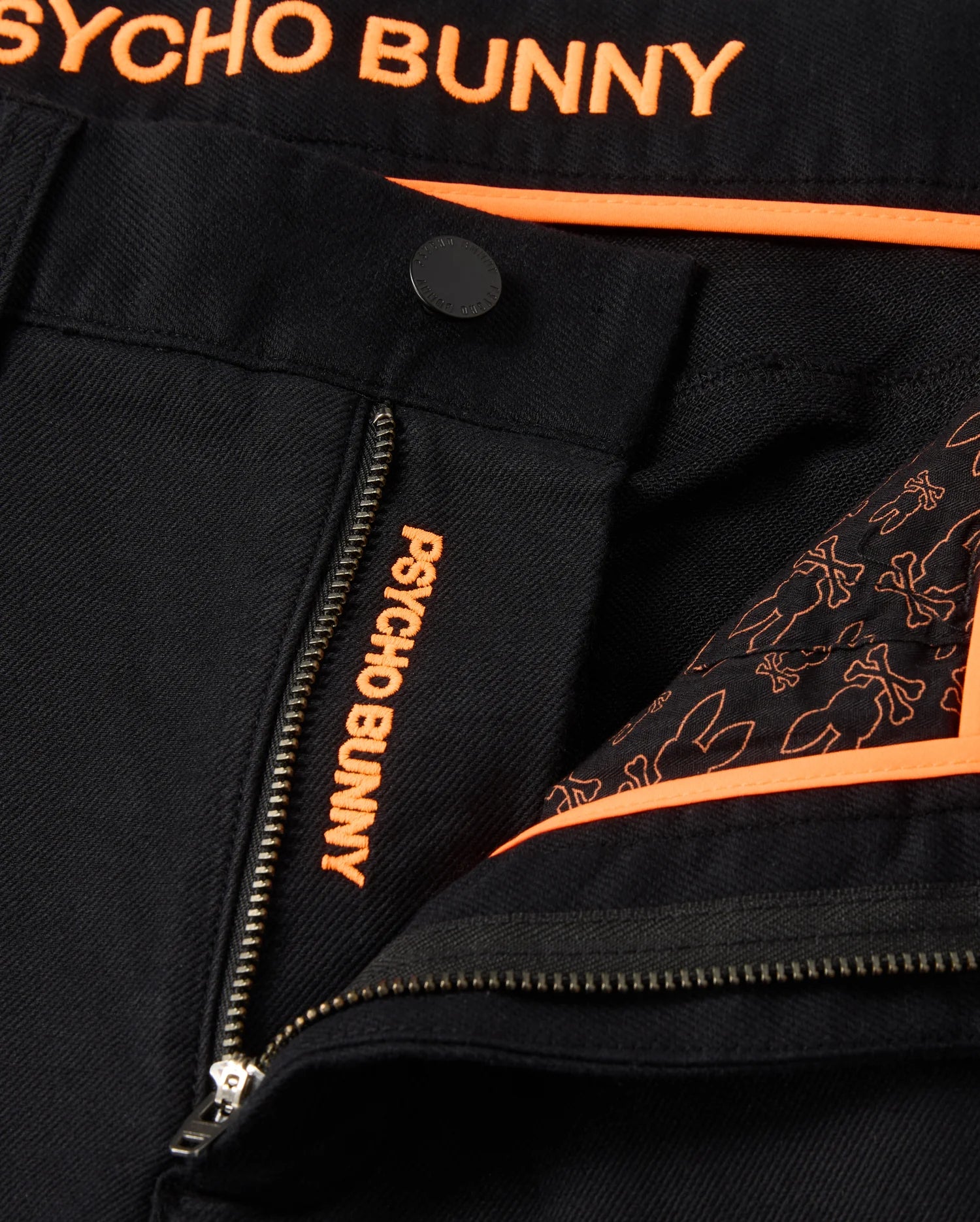 Close-up of the black fabric on the MENS HAYS TWILL PANT - B6P894D200 by Psycho Bunny, featuring orange text "PSYCHO BUNNY" on a zipper and embroidered detail. The lining, made from a cotton-blend twill, showcases a pattern of bunny outlines in orange. Orange piping along the garment's edge adds to its ultimate comfort.