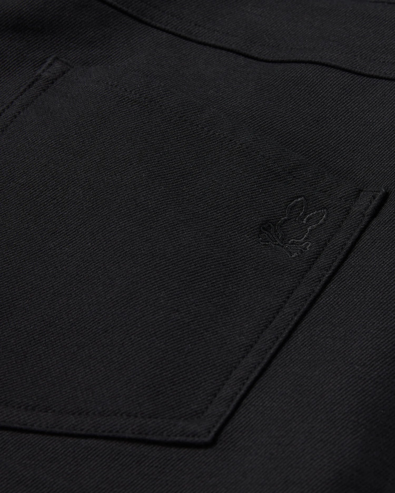 Close-up of the Psycho Bunny MENS HAYS TWILL PANT in black, made from a cotton-blend twill. It features a pocket adorned with an embroidered rabbit logo in the corner. The fabric has a textured appearance and provides ultimate comfort with its 4-way stretch design.