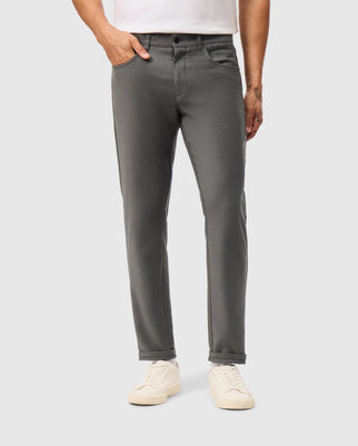 A person wearing Psycho Bunny's dark gray MENS HAYS TWILL PANT - B6P894D200, with hands in pockets, paired with a white shirt. The slim-fit pants harmonize perfectly with white sneakers against a plain light gray background.