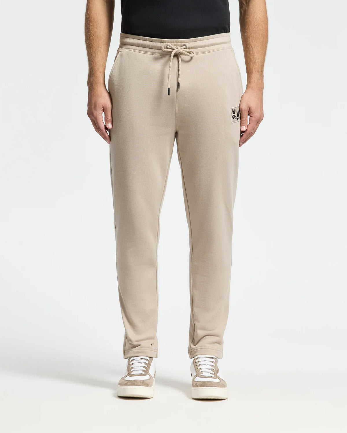 A person wearing Psycho Bunny's beige MENS MONROE TWILL PATCH FLEECE SWEATPANT featuring a drawstring and small embroidered logo on the left pocket, paired with a black shirt and white sneakers, stands against a plain white background.
