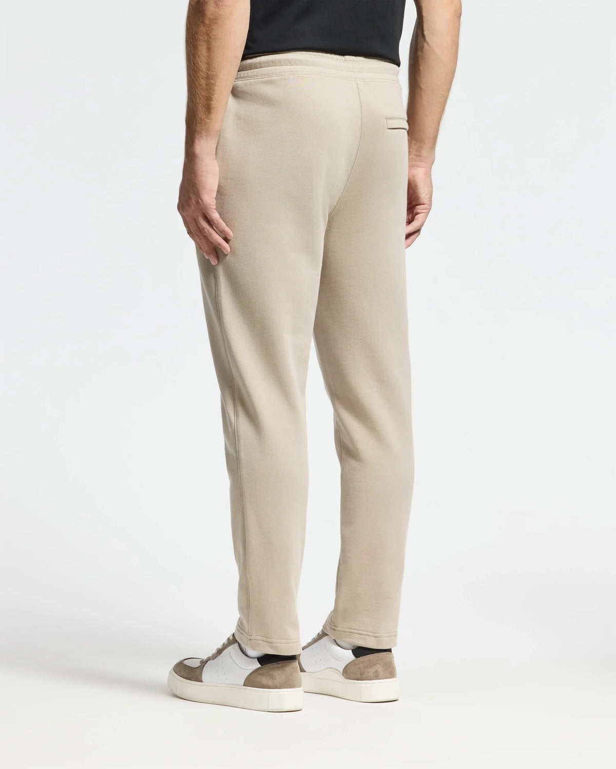 A person is seen from behind wearing beige Monroe twill patch fleece sweatpants by Psycho Bunny and a black shirt. The look is complemented by white sneakers with brown accents against a plain white background, emphasizing the casual outfit.